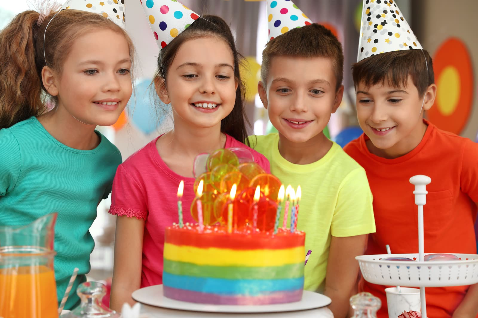 Four children in party hats looking at a rainbow birthday cake with lit candles
