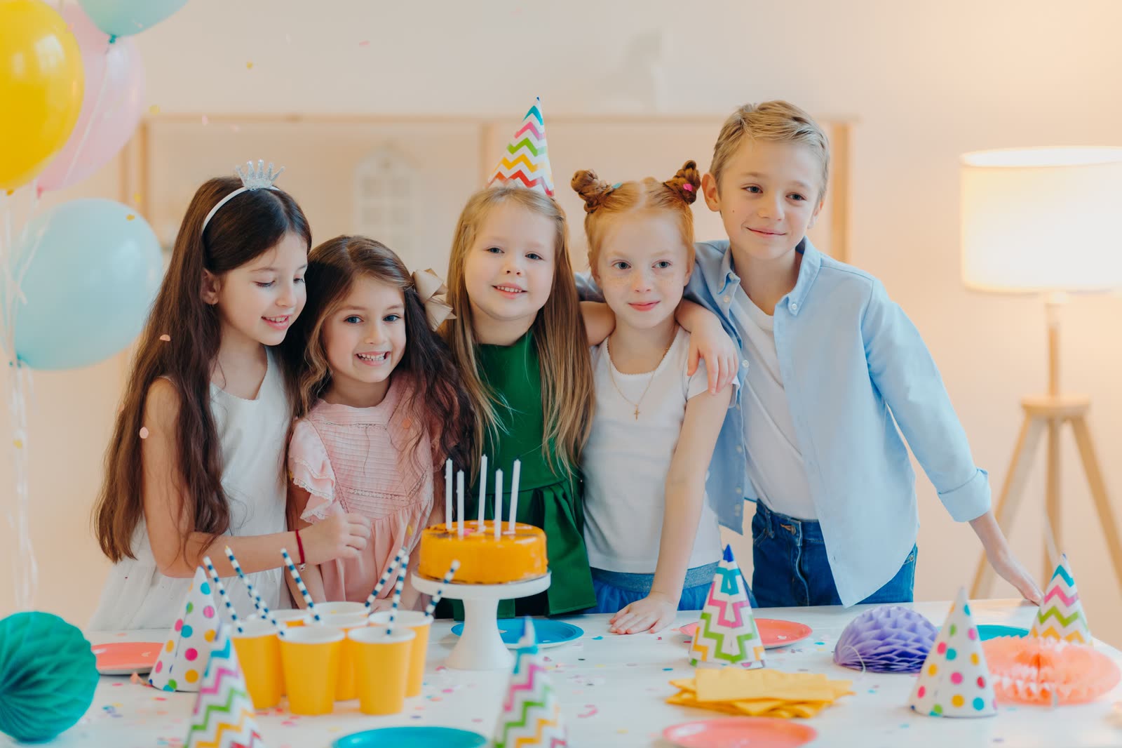 Five friends at a birthday party table with a cake, party hats and balloons